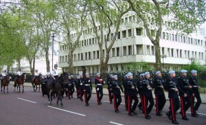 A group of uniformed soldiers is marching in formation on a street lined with trees. Some soldiers are on horseback, while others are walking. The background features a large, multi-story building with numerous windows. The scene appears to be part of a formal parade or procession.