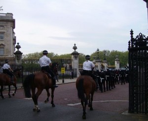 The image shows a group of mounted police officers riding horses, leading a procession of uniformed officers on foot. They are moving through a gated area with ornate iron gates, and there is a large building visible in the background. The scene appears to be part of a formal event or ceremony.