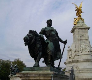 The image shows a bronze statue of a man standing beside a lion, both mounted on a pedestal. In the background, there is a tall, ornate monument topped with a golden statue of a winged figure. The sky is overcast, and there are trees visible in the distance.