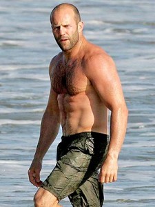 A muscular man with a shaved head is standing in shallow water at the beach. He is shirtless and wearing dark swim trunks. The background shows the ocean with gentle waves.