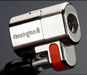 The image shows a close-up of a Kensington laptop lock. The lock is metallic with a shiny finish and has the brand name "Kensington" engraved on it. There is a red button or switch on the side of the lock. The lock is designed to secure laptops and other devices.