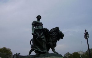 A large bronze statue depicting a woman standing beside a lion. The woman is dressed in classical attire and holds a sickle in her right hand. The lion stands to her right, appearing strong and majestic. The statue is set against a cloudy sky, with some trees and a lamp post visible in the background.