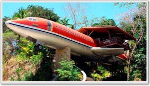 The image shows a repurposed airplane that has been converted into a unique building or structure. The airplane is mounted on a stone platform and surrounded by lush greenery and trees. The structure appears to be integrated into a hillside, with a wooden deck or patio extending from the side of the airplane. The airplane is painted red and white, and the setting suggests it might be used as a hotel, restaurant, or some other type of accommodation or attraction.
