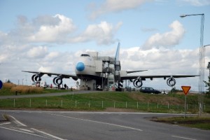 The image shows a large airplane that appears to be repurposed as a building or structure. The airplane is situated on a grassy area near a road, with a staircase and other structural elements attached to it. There are a few cars parked nearby, and the sky is partly cloudy.