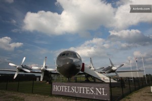 The image shows a large airplane displayed outdoors on a grassy area, surrounded by a black fence. The airplane has four propellers and is positioned facing the camera. In front of the airplane, there is a sign that reads "HOTELSUITES.NL." The sky above is partly cloudy, and there is a small building visible in the background. The image also has a watermark in the top right corner that says "Airbnb.com."