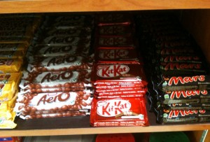 The image shows a shelf stocked with various chocolate bars. From left to right, there are Aero bars, KitKat bars, and Mars bars. The Aero bars are in white and brown packaging, the KitKat bars are in red packaging, and the Mars bars are in black packaging.