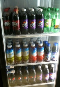 The image shows a refrigerated display case filled with various bottled beverages. The top shelf contains bottles of Dr. Pepper, Crush grape soda, and 7UP. The middle shelf has bottles of flavored water, including lemon and berry flavors. The bottom shelf features bottles of Pepsi and more Crush grape soda.