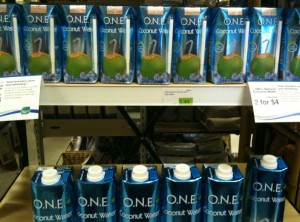 The image shows a store shelf stocked with multiple cartons of O.N.E. Coconut Water. The cartons are blue and feature an image of a green coconut with a straw. There are two rows of the cartons, with a sign indicating a sale price of "2 for $4."