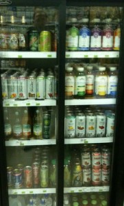 The image shows a refrigerated display case filled with various beverages. The drinks are organized on multiple shelves behind glass doors. The assortment includes cans and bottles of different sizes and colors, featuring a variety of brands and flavors. The labels on the drinks indicate a mix of sodas, juices, and other bottled beverages.