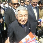 A smiling elderly man with gray hair, dressed in a dark suit and tie, is holding a bouquet of flowers and a small Canadian flag. He is surrounded by several people, some of whom are wearing sunglasses and suits. The group appears to be outdoors, and there are more people and greenery visible in the background.