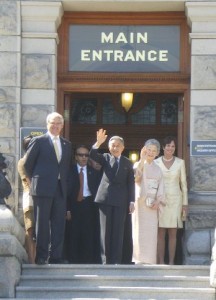 A group of people standing on the steps in front of a building with a sign that reads "Main Entrance." One person in the center is waving. The group includes both men and women, dressed in formal attire. The building has a stone facade and wooden doors.