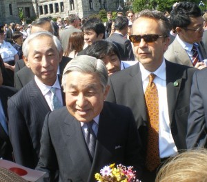 A group of people, including an elderly man in a dark suit holding a bouquet of flowers, are gathered outdoors. The elderly man is surrounded by other men in suits, one of whom is wearing sunglasses. There are more people in the background, and the setting appears to be a formal event or gathering.