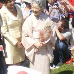 A woman in a traditional Japanese kimono is holding a bouquet of flowers and standing next to another woman in a light-colored suit. They are outdoors, surrounded by a crowd of people, some of whom are taking photos and waving Japanese flags. The atmosphere appears to be celebratory.