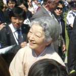 An elderly woman with gray hair, wearing a light-colored traditional outfit, is smiling and interacting with a crowd of people. The crowd includes men in suits and ties, some of whom appear to be security personnel, and a police officer in uniform. The background shows a sunny outdoor setting with trees and more people, some of whom are taking photos.