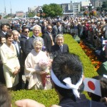 A large crowd is gathered outdoors, with many people holding Japanese flags. In the foreground, a group of dignitaries, including a woman in a traditional kimono holding a bouquet of flowers, are posing for a photo. The background shows a bustling scene with more people, buildings, and greenery. The atmosphere appears to be celebratory and formal.