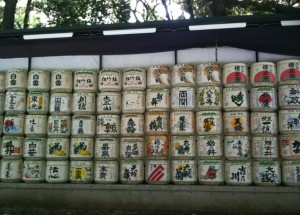 The image shows a wall of stacked traditional Japanese sake barrels, known as "kazaridaru," which are often seen at Shinto shrines. The barrels are decorated with various colorful labels and Japanese characters. The barrels are arranged in a grid pattern, and the wall is located outdoors, with trees visible in the background.