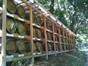 The image shows a series of wooden barrels stacked in a wooden frame structure outdoors. The barrels are arranged in multiple rows and columns, and each barrel has a plaque with text on it. The structure is situated in a wooded area with trees and greenery surrounding it. Sunlight filters through the trees, casting dappled shadows on the barrels and the ground.