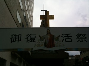 The image shows a sign with Chinese characters and an illustration of Jesus Christ with his arms open. Above the sign, there is a large cross. The background includes a tall building and a cloudy sky.