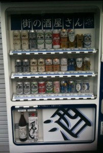 The image shows a vending machine stocked with various alcoholic beverages. The machine has multiple rows of cans and bottles, including beer and sake. The top three rows contain cans of different brands and types, while the bottom row has larger bottles. The vending machine has Japanese text on it, indicating that it is likely located in Japan.