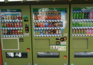 The image shows three green vending machines lined up side by side. Each vending machine is stocked with a variety of beverages, including bottles and cans of different colors and sizes. The machines have selection buttons below each drink, and there are payment slots and dispensing areas at the bottom. The branding "ITO EN" is visible at the top of each machine.