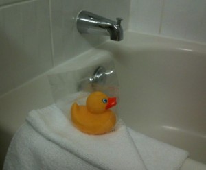 The image shows a yellow rubber duck with an orange beak placed on a white towel. The towel is draped over the edge of a bathtub, and a silver faucet is visible in the background. The rubber duck is in a clear plastic bag.