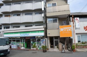 The image shows a street view of a small commercial area. On the left, there is a FamilyMart convenience store with a green and white sign. To the right of the FamilyMart, there is a narrow building with a large orange sign that reads "Caro Hair Design." The buildings have multiple stories, and there is a white truck parked in front of the FamilyMart. The area appears to be clean and well-maintained, with some potted plants and bicycles visible near the entrances.