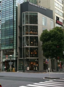 The image shows a multi-story building with a sign at the top that reads "FRESHNESS BURGER." The building has large glass windows on each floor, allowing a view of the interior. It is situated on a street corner in an urban area, with other tall buildings and a tree visible nearby. The street in front of the building has a pedestrian crosswalk.