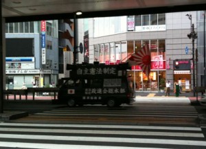 A black van with Japanese text and a large Japanese flag on top is driving through a city street. The van is partially obscured by shadows from an overhead structure. In the background, there are modern buildings with glass windows and various storefronts, including a brightly lit shop with a red and white sign. The street has a pedestrian crosswalk in the foreground.