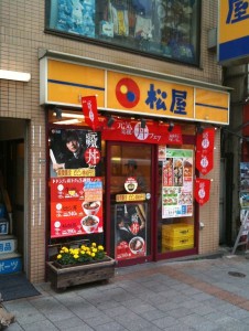 The image shows the exterior of a Japanese restaurant. The storefront has a yellow and blue sign with Japanese characters and a red and blue circular logo. The entrance is decorated with red banners and posters displaying menu items and promotional images. There is a small flower box with yellow flowers on the left side of the entrance. The restaurant appears to be located on a city street with a tiled sidewalk in front.