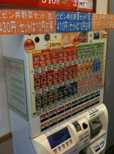 The image shows a Japanese vending machine used for ordering food. It has numerous buttons with pictures and prices of different meal options. The machine has a display screen and a slot for inserting money. There are also signs with Japanese text above the machine, likely providing additional information or promotions.