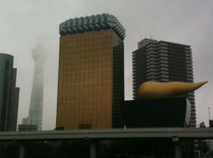 The image shows a cityscape with several notable buildings. In the foreground, there is a large golden building with a unique, wavy roof design. To the right of this building, there is a black structure with a large, golden, flame-like sculpture on top. In the background, there is a tall, slender tower partially obscured by fog or clouds. Other modern buildings are also visible around these structures. The overall scene appears to be taken on a cloudy or foggy day.