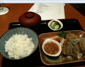 A meal is presented on a tray, consisting of a bowl of white rice, a small bowl of soup with a lid, a plate with fried food and a dipping sauce, and a small dish of sliced cucumbers. A menu is visible in the background.