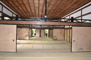 The image shows the interior of a traditional Japanese room with tatami mat flooring. The room features sliding doors (fusuma) with simple, natural designs. The ceiling is made of wooden beams, and there is a visible wooden framework supporting the structure. In the background, there are more sliding doors leading to other rooms or areas, and some greenery can be seen through the open doors. The overall atmosphere is serene and minimalist, typical of traditional Japanese architecture.