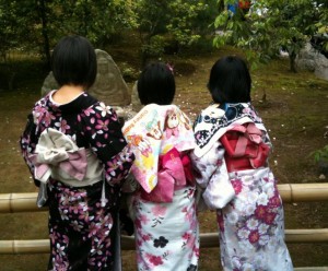 Three people are standing with their backs to the camera, wearing colorful kimonos with intricate patterns. They are in an outdoor setting with greenery and trees in the background. The kimonos have different floral designs and are tied with obi sashes. The scene appears to be serene and traditional.