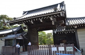 The image shows a traditional Japanese gate, likely the entrance to a historical or cultural site. The gate features a large wooden structure with a tiled roof and intricate architectural details. There are two security guards standing near the gate, and a wooden fence partially blocks the entrance. Trees and greenery are visible in the background, suggesting the gate leads to a garden or park area.