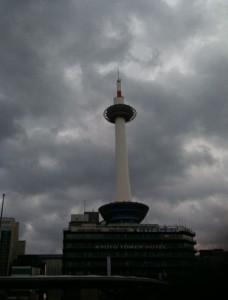 The image shows a tall observation tower with a circular observation deck near the top, set against a backdrop of dark, cloudy skies. The tower is situated above a multi-story building, and the overall scene appears to be in an urban area. The atmosphere is moody due to the overcast weather.