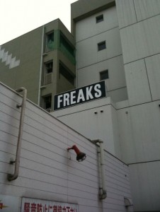 The image shows a multi-story building with a sign on the roof that reads "FREAKS" in large, bold letters. The building has a modern design with a mix of white and gray exterior walls. There is a red security camera mounted on the side of the building. The lower part of the image includes a sign with Japanese text.