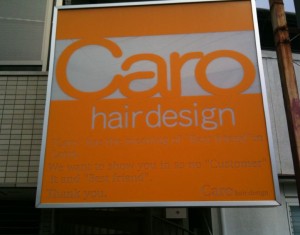 The image shows a sign for a hair salon named "Caro hair design." The sign has an orange background with white text. The main text reads "Caro hair design" in large letters. Below that, there is smaller text that is partially readable, mentioning something about "Customer" and "Best friend." The sign is mounted on a building exterior.