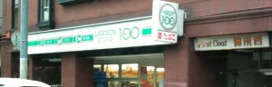 The image shows the exterior of a convenience store with a sign that reads "Lawson Store 100." The store has a green and white color scheme. There is another sign on the right side of the image with Japanese characters, indicating another business next to the convenience store. The building appears to be made of brick.