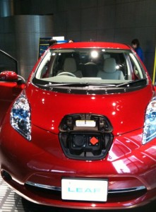 A red electric car is shown from the front with its charging port open. The car has a license plate that reads "LEAF." The background includes a person and some indoor elements, suggesting the car is displayed indoors, possibly at an auto show or dealership.