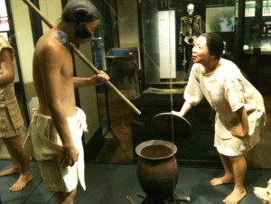 The image shows a museum exhibit featuring two life-sized mannequins dressed in traditional clothing. One mannequin, appearing to be male, is holding a long stick and standing next to a large pot. The other mannequin, appearing to be female, is leaning over the pot, seemingly engaged in a conversation or activity related to the pot. The background includes various museum displays and a skeleton model.