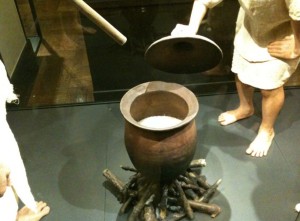 The image shows a large clay pot placed over a fire made of logs. The pot is being opened by a person, revealing its contents. The scene appears to be a historical or cultural display, possibly in a museum, depicting traditional cooking methods. The individuals in the image are dressed in simple, historical clothing.