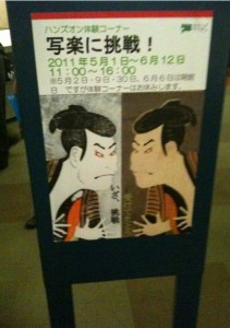 The image shows a poster on a stand. The poster features two traditional Japanese illustrations of men facing each other, both with stern expressions. The text on the poster is in Japanese and includes dates and times, specifically mentioning "2011年5月1日～6月12日" and "11:00～16:00". The poster appears to be advertising an event or exhibition.