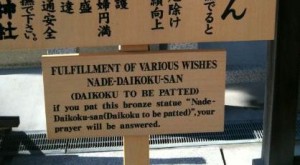 The image shows a wooden sign with text in both English and Japanese. The English text reads: "FULFILLMENT OF VARIOUS WISHES NADE-DAIKOKU-SAN (DAIKOKU TO BE PATTED) if you pat this bronze statue 'Nade-Daikoku-san (Daikoku to be patted)', your prayer will be answered." The sign is placed outdoors, and part of another sign with Japanese text is visible above it.