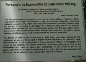 The image shows a sign with text explaining the provenance of Bourgogne wine for consecration at Meiji Jingu. The text describes the historical context of the Meiji period, emphasizing the blending of Japanese spirit and Western knowledge. It mentions the role of Emperor Meiji in promoting modernization while preserving Japanese traditions. The sign also details the donation of wine barrels from Bourgogne, France, facilitated by various individuals and organizations, as a symbol of friendship between France and Japan. The text concludes with a note of gratitude and a hope for continued peace and amity between the two nations.