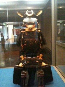 The image shows a traditional Japanese samurai armor set displayed in a glass case. The armor is black with intricate detailing and features a helmet with a prominent crescent moon crest. The armor is positioned in a seated posture, and the display is located in a museum or exhibition setting.