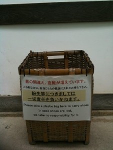 A woven basket is placed against a wall. Attached to the front of the basket is a sign with text in both Japanese and English. The English text reads: "Please take a plastic bag here to carry shoes. In case shoes are lost, we take no responsibility for it." The basket appears to be intended for visitors to place their shoes in, possibly at the entrance of a building.