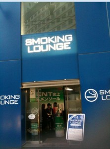 The image shows the entrance to a smoking lounge. The entrance is marked by a large blue sign with white text that reads "SMOKING LOUNGE." There is a smaller sign to the right of the entrance with a smoking symbol and the same text. Inside, there are people visible through the glass doors, and there is a green advertisement or sign in the background. There is also a stand with a signboard in front of the entrance.