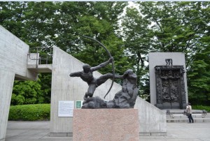 The image shows a statue of a person drawing a bow, mounted on a stone pedestal. The statue is part of an outdoor display with concrete structures and a large, intricate relief in the background. The area is surrounded by lush green trees, and a person can be seen walking in the background.