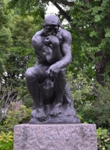 The image shows a statue of a man sitting on a rock with his chin resting on his hand, appearing to be deep in thought. The statue is surrounded by greenery, including trees and bushes. The statue is mounted on a stone pedestal.